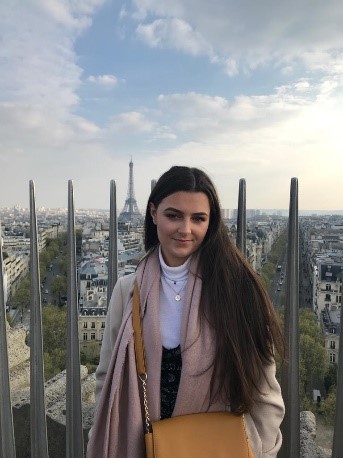 Abigail, a young white woman with long brown hair, standing with her back to a city view of the Paris skyline