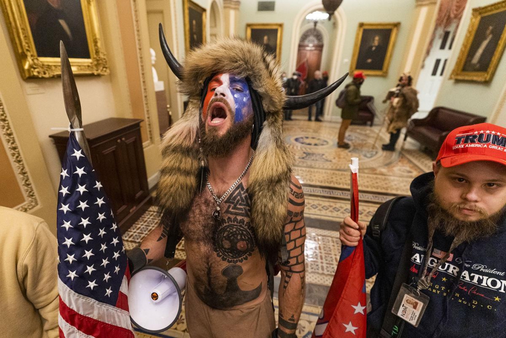 Two male protestors from the attempted coup in January. The one in the centre is shirtless, showing tribal and nordic influenced tattoos. He is carrying a spear (with an American flag tied to it) and a megaphone. He has the US flag painted on his face, and is wearing an animal fur hat with horns. The other man is also carrying a flag, and has a beard. He is wearing a hoodied and a "trump" hat. They are both standing in a hall in the US Senate building. 