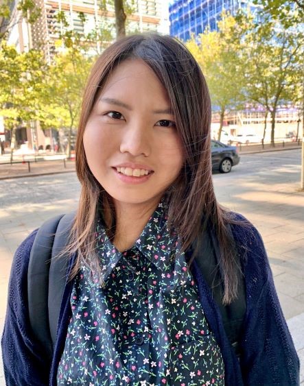 Mirai is a young Japanese woman, with long brown hair. She is wearing a shirt with a flower pattern and a cardigan. She is standing outside in a urban area, with small trees in the backgroun