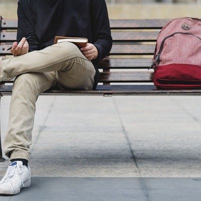 male-student-sitting-on-bench-with-backpack-pen-and-book